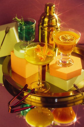 A stylish arrangement of three cocktail glasses, each filled with different vibrant drinks, placed on a reflective golden tray. One drink is green with rosemary, another yellow adorned with an orange flower, and the third orange topped with a citrus slice. A shiny cocktail shaker stands in the background, creating a luxurious and festive atmosphere.