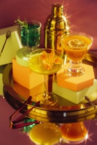 A stylish arrangement of three cocktail glasses, each filled with different vibrant drinks, placed on a reflective golden tray. One drink is green with rosemary, another yellow adorned with an orange flower, and the third orange topped with a citrus slice. A shiny cocktail shaker stands in the background, creating a luxurious and festive atmosphere.