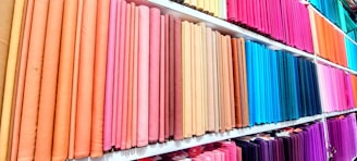 Various brightly colored fabric rolls are neatly arranged on shelves in a fabric store. The fabrics range from pastel pinks and peaches to vibrant blues, purples, and greens.
