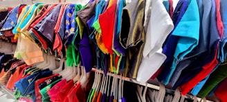 A vibrant assortment of clothing hangs on white hangers. The clothes feature a variety of colors and patterns, including reds, blues, greens, purples, and a mix of textures and fabrics. The garments appear to be neatly arranged on a rack in what seems to be a retail setting.