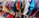 A vibrant assortment of clothing hangs on white hangers. The clothes feature a variety of colors and patterns, including reds, blues, greens, purples, and a mix of textures and fabrics. The garments appear to be neatly arranged on a rack in what seems to be a retail setting.