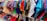 A vibrant assortment of clothing hangs on white hangers. The clothes feature a variety of colors and patterns, including reds, blues, greens, purples, and a mix of textures and fabrics. The garments appear to be neatly arranged on a rack in what seems to be a retail setting.