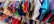 A vibrant assortment of clothing hangs on white hangers. The clothes feature a variety of colors and patterns, including reds, blues, greens, purples, and a mix of textures and fabrics. The garments appear to be neatly arranged on a rack in what seems to be a retail setting.