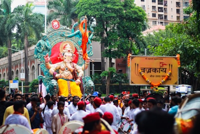 A large, elaborately decorated statue of Lord Ganesha is being paraded through a street lined with trees and buildings. The statue is adorned with vivid colors, particularly red, yellow, and teal. A crowd of people, some dressed in traditional clothing, can be seen surrounding the statue, participating in the procession. Many are wearing white clothing and red caps, and a sign with text is being carried by the crowd.
