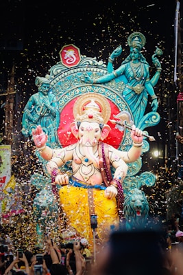 A large, colorful statue of a deity adorned with intricate patterns and bright yellow garments stands prominently in a festive setting. Surrounding the statue are smaller figures and elaborate decorations, with confetti falling from above. There are many people gathered around, capturing the moment with their phones.