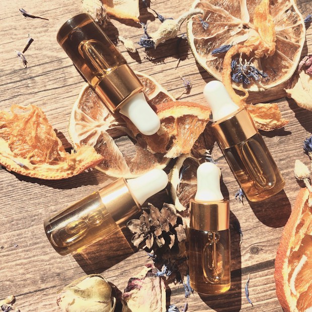 A flat lay composition featuring several glass dropper bottles with white droppers and gold accents, surrounded by dried citrus slices, pinecones, and small flowers. The setup is placed on a wooden surface, creating a warm, rustic aesthetic.
