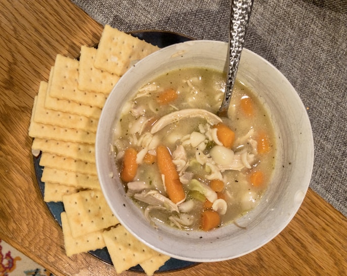 A bowl of chicken noodle soup is placed on a wooden table, accompanied by a stack of square crackers. The soup contains pieces of chicken, carrots, and pasta in a light broth, with a spoon resting inside the bowl. The setting suggests a warm and comforting meal.