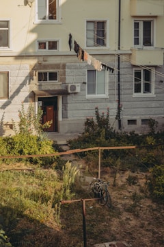 An old apartment building with a yellow and gray facade. Windows have some of the curtains drawn and laundry is hanging on a line outside. There is a small, overgrown garden in the foreground with a bicycle leaning against a wooden structure. Sunlight casts shadows across the building and garden.