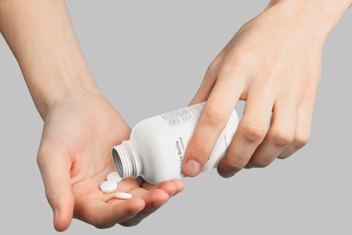 Hands are seen holding and pouring pills from a white bottle into an open palm against a neutral background.