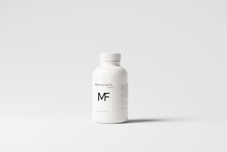 A white plastic bottle with a screw top, labeled 'Medicine Bottle mockup' and the letters 'MF' in bold. The bottle is placed against a plain, light gray background, with some small text details visible on the right side.