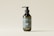 A minimalist design of a dark green cosmetic bottle with a pump dispenser, placed against a light beige background. The bottle has a label with white text and a simple logo.