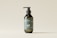 A minimalist design of a dark green cosmetic bottle with a pump dispenser, placed against a light beige background. The bottle has a label with white text and a simple logo.