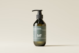 A minimalist design of a dark green cosmetic bottle with a pump dispenser, placed against a light beige background. The bottle has a label with white text and a simple logo.
