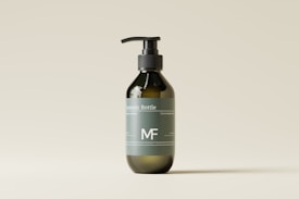 A minimalist design of a dark green cosmetic bottle with a pump dispenser, placed against a light beige background. The bottle has a label with white text and a simple logo.