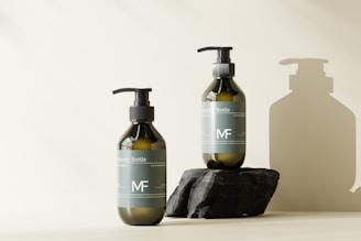 Two green-tinted pump bottles are displayed on a surface against a light background. One bottle stands directly on the surface, while the other is elevated on a dark, textured stone. Both bottles have a minimalist design with white labels and are casting soft shadows.