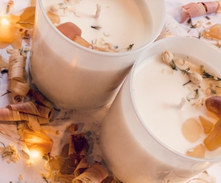 Two creamy white candles in glass holders are surrounded by small, rustic wood shavings and delicate flowers. Ambient warm lighting casts a soft glow on the scene, contributing to a cozy atmosphere.