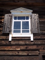 A rustic wooden window with weathered shutters is part of a log cabin exterior. The wood appears aged and textured, with a slightly distressed appearance. The window has a white frame with a triangular pediment on top, showing signs of rust.