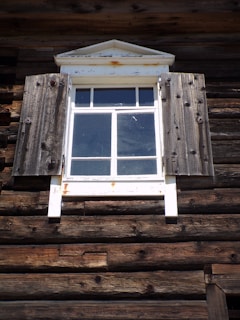 A rustic wooden window with weathered shutters is part of a log cabin exterior. The wood appears aged and textured, with a slightly distressed appearance. The window has a white frame with a triangular pediment on top, showing signs of rust.