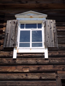 A rustic wooden window with weathered shutters is part of a log cabin exterior. The wood appears aged and textured, with a slightly distressed appearance. The window has a white frame with a triangular pediment on top, showing signs of rust.