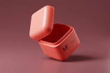 A pink cube-shaped container with a lid slightly ajar, placed against a similarly colored background. The container features text and the letters MF on its side.