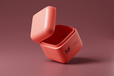 A pink cube-shaped container with a lid slightly ajar, placed against a similarly colored background. The container features text and the letters MF on its side.