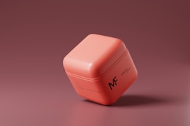 A small, cubic container with a smooth, matte finish is featured. The container is a soft coral color and has minimalistic black text that reads 'MF' and 'cube jar' on the side. The background is a gradient of darker shades of coral, giving the image a modern and sleek appearance.