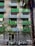 An urban apartment building featuring modern green balconies, with several small trees in front. A row of parked scooters lines the sidewalk, and a tall palm tree is visible on the right. A striped flag is displayed on one of the balconies.