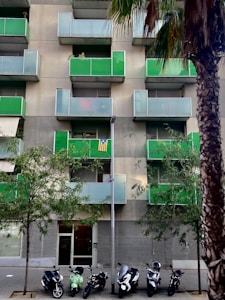 An urban apartment building featuring modern green balconies, with several small trees in front. A row of parked scooters lines the sidewalk, and a tall palm tree is visible on the right. A striped flag is displayed on one of the balconies.