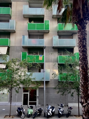 An urban apartment building featuring modern green balconies, with several small trees in front. A row of parked scooters lines the sidewalk, and a tall palm tree is visible on the right. A striped flag is displayed on one of the balconies.