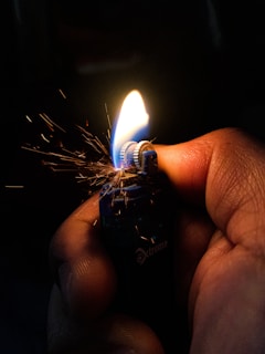 A hand is holding a lighter, with a flame burning brightly. Sparks are visible around the ignition wheel, creating a dynamic and energetic effect. The background is dark, highlighting the flame and the hand holding the lighter.