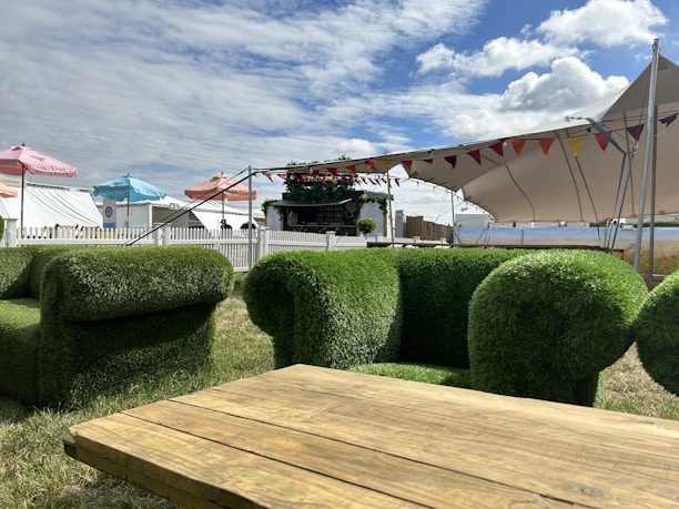 The scene features outdoor furniture designed to resemble grass, including chairs and a table. A large tent with colorful flags provides shade in the background, while several umbrellas add pops of blue, pink, and red. A white picket fence encloses the area, which appears to be part of a festival or event setup, complete with a small bar or kiosk decorated with plants.