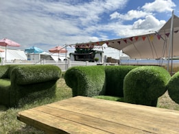 The scene features outdoor furniture designed to resemble grass, including chairs and a table. A large tent with colorful flags provides shade in the background, while several umbrellas add pops of blue, pink, and red. A white picket fence encloses the area, which appears to be part of a festival or event setup, complete with a small bar or kiosk decorated with plants.