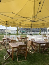 A shaded outdoor seating area with wooden tables and chairs under a large yellow tent. The seating area is situated on a grassy field, enclosed by a white picket fence. In the background, there are buildings and additional tents, possibly indicating a fair or event location.
