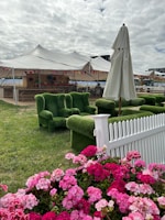 An outdoor setting featuring a fenced garden area with vibrant pink flowers in the foreground. Behind the flowers, there are green grass-covered armchairs and a white umbrella. A large tent with an open side is visible in the background, with colorful bunting hanging from it. The sky is overcast with grey clouds.