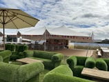 A spacious outdoor setting featuring green grass-covered sofas arranged around wooden coffee tables, under large umbrellas. In the background, there is a large white event tent decorated with colorful bunting, with a bar area and seating.