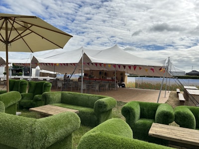 A spacious outdoor setting featuring green grass-covered sofas arranged around wooden coffee tables, under large umbrellas. In the background, there is a large white event tent decorated with colorful bunting, with a bar area and seating.
