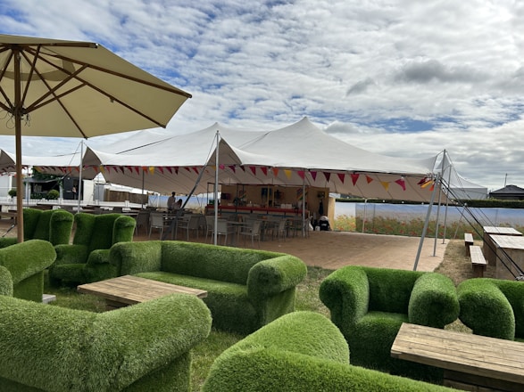 A vibrant outdoor event under a large cream-colored stretch tent with wooden furniture and soft lighting.