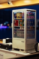 A beverage display fridge is filled with various drinks, including Pepsi, Coca-Cola, Mirinda, Lipton, and water bottles. It sits on a countertop with a cash register and a kettle nearby. The background is blurred with bokeh lights, indicating an indoor setting.
