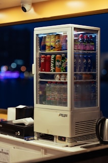 A beverage display fridge is filled with various drinks, including Pepsi, Coca-Cola, Mirinda, Lipton, and water bottles. It sits on a countertop with a cash register and a kettle nearby. The background is blurred with bokeh lights, indicating an indoor setting.