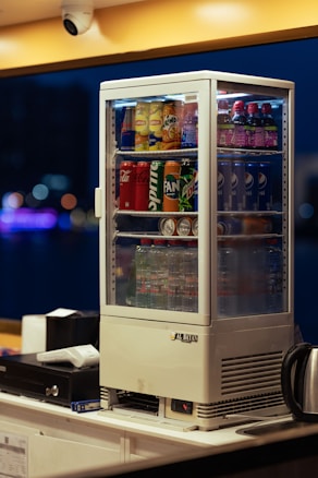 A beverage display fridge is filled with various drinks, including Pepsi, Coca-Cola, Mirinda, Lipton, and water bottles. It sits on a countertop with a cash register and a kettle nearby. The background is blurred with bokeh lights, indicating an indoor setting.