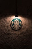 A logo featuring a mermaid with two tails is illuminated by a single overhead light against a textured, dark wall.