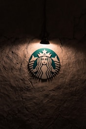 A logo featuring a mermaid with two tails is illuminated by a single overhead light against a textured, dark wall.
