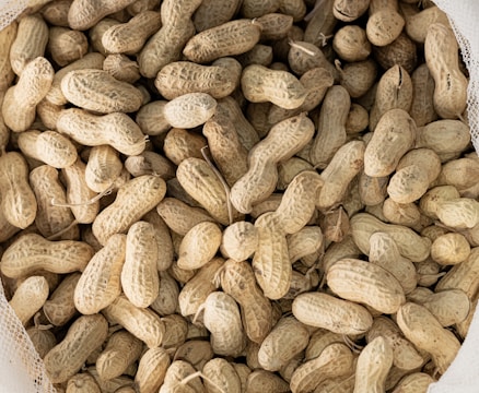 A pile of unshelled peanuts filling the frame, with a variety of sizes and organic shapes. The peanuts have a textured, rough shell and an earthy hue. The image captures the dry, fibrous appearance typical of raw peanuts.