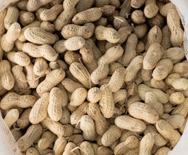 A pile of unshelled peanuts filling the frame, with a variety of sizes and organic shapes. The peanuts have a textured, rough shell and an earthy hue. The image captures the dry, fibrous appearance typical of raw peanuts.