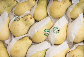 Several pears are arranged together, each wrapped in white paper and labeled with green stickers. The fruits have a pale yellow color and are stacked closely in rows.