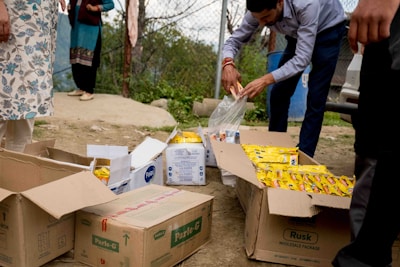 A person is sorting through packages of snacks on the ground, surrounded by several open cardboard boxes containing items like biscuits and rusk. Other individuals are partially visible, standing nearby on a dirt area near a fence, with trees and hills in the background.