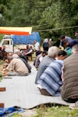 A group of people gathering outdoors seated on large mats, possibly participating in a communal meal or event. The setting appears to be in a forested or rural area with trees surrounding the scene. There is a vehicle in the background with a blue tarpaulin, and some people are standing near it. The overall atmosphere is casual and communal.