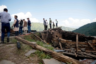 A group of people stands on a grassy slope, observing a partially collapsed stone structure. The area is surrounded by lush green mountains and a clear blue sky with clouds. Some individuals are dressed in traditional attire while others wear casual clothing.