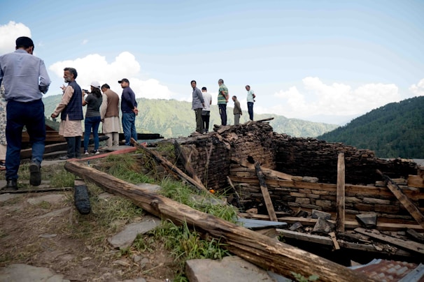 A group of people stands on a grassy slope, observing a partially collapsed stone structure. The area is surrounded by lush green mountains and a clear blue sky with clouds. Some individuals are dressed in traditional attire while others wear casual clothing.