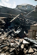 Charred and collapsed wooden beams and debris are scattered across a pile, with a partially damaged roof visible in the background. The scene suggests destruction, likely from a fire.